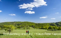 Zwei Wanderer laufen bei blauem Himmel über eine grüne Wiese